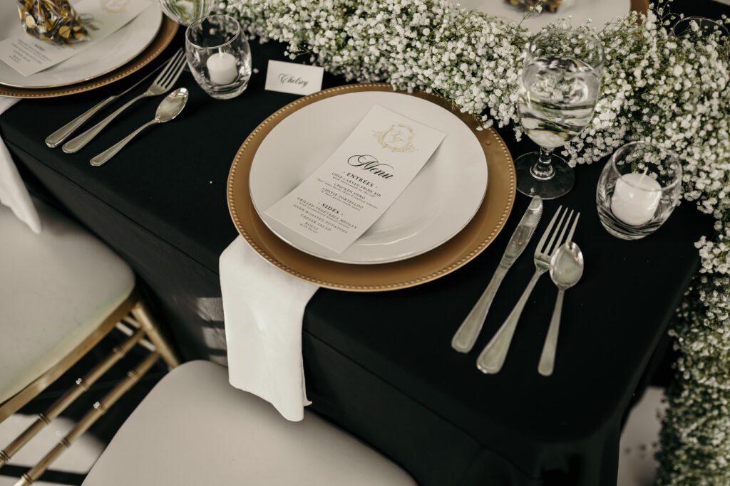 Wedding reception tables cape with a baby's breath table runner, custom signage, silver flatware, and gold chargers.