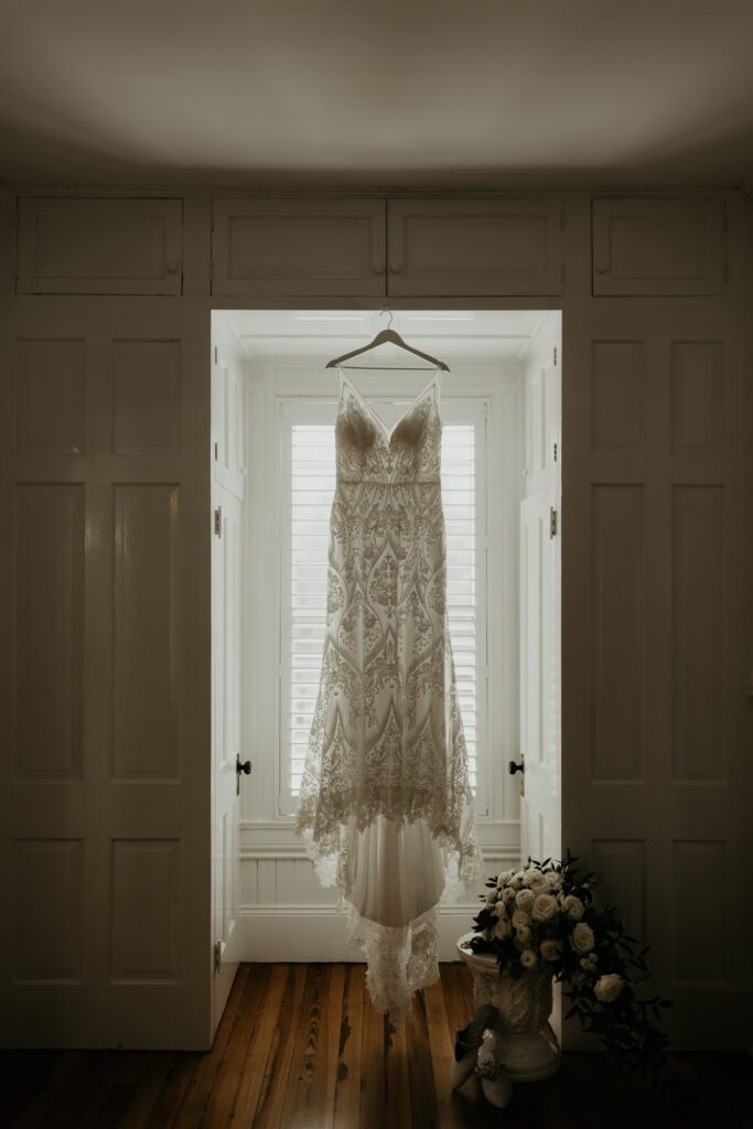 Moody wedding dress hanging in the window of the wedding venue with the bride's bouquet and shoes.