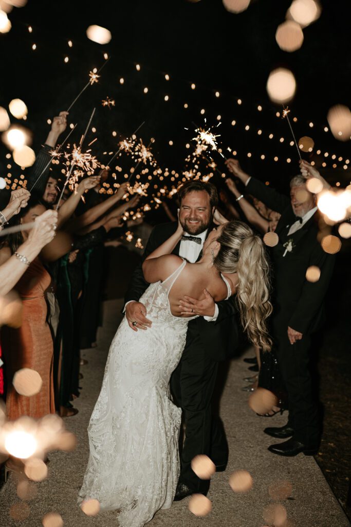 documentary style photo of bride with ponytail and a deep v neck wedding dress going in for a dip kiss at their sparkler exit.