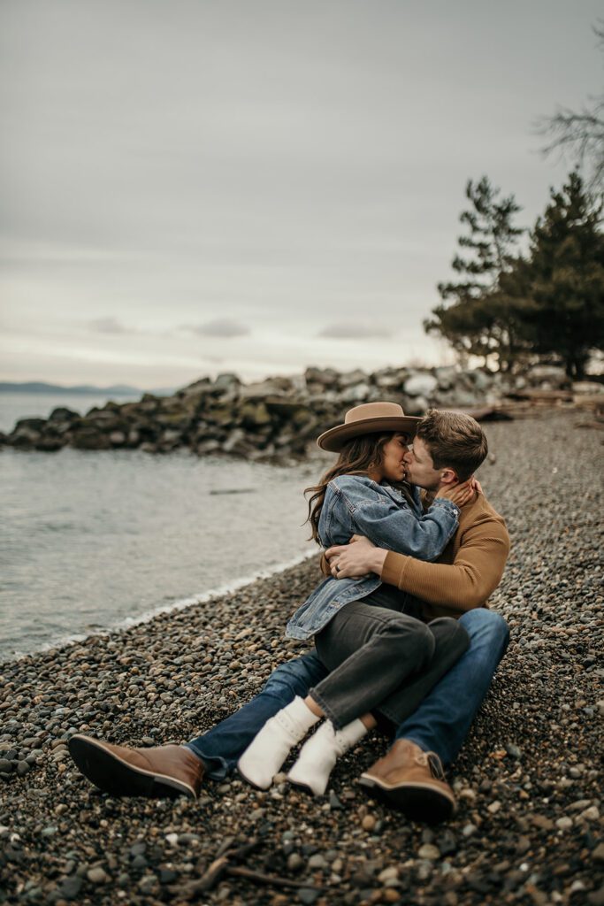 portrait of a couple kissing on the beach of the puget sound with evergreen trees visible in the background.